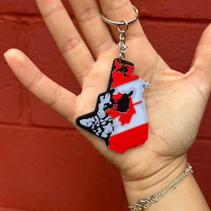 May include: A keychain featuring a map of Canada with a red and white flag design. The keychain has a silver keyring and chain. The map includes black and white details. The keychain is held in a hand against a red background.