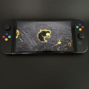 May include: A black handheld gaming console with a colourful button layout. The screen displays a black and gold background with a stylised black panther logo.