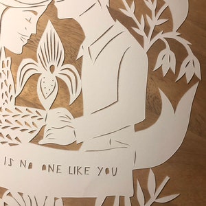 May include: A white paper cut-out artwork featuring a couple, flowers, and foliage. The text "IS NO ONE LIKE YOU" is cut into a banner. The artwork is set against a wooden background.