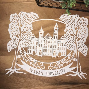May include: White paper cut-out design of a building with two trees on either side. The building has a large central tower and smaller towers on either side. The text "Auburn University" is written below the building.