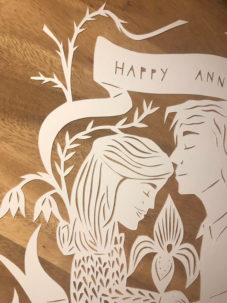May include: A white paper cut-out art piece featuring a couple embracing, with a floral design and the words "HAPPY ANN" cut into a banner. The artwork is set against a wooden background.
