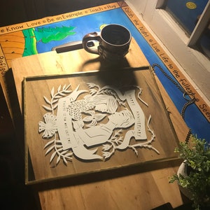 May include: A framed paper cut-out with a wood backing. The white cut-out features a couple embracing, floral elements, and the words "HAPPY ANNIVERSARY" and "THERE IS NO ONE LIKE YOU". A brown mug sits on a wooden surface.