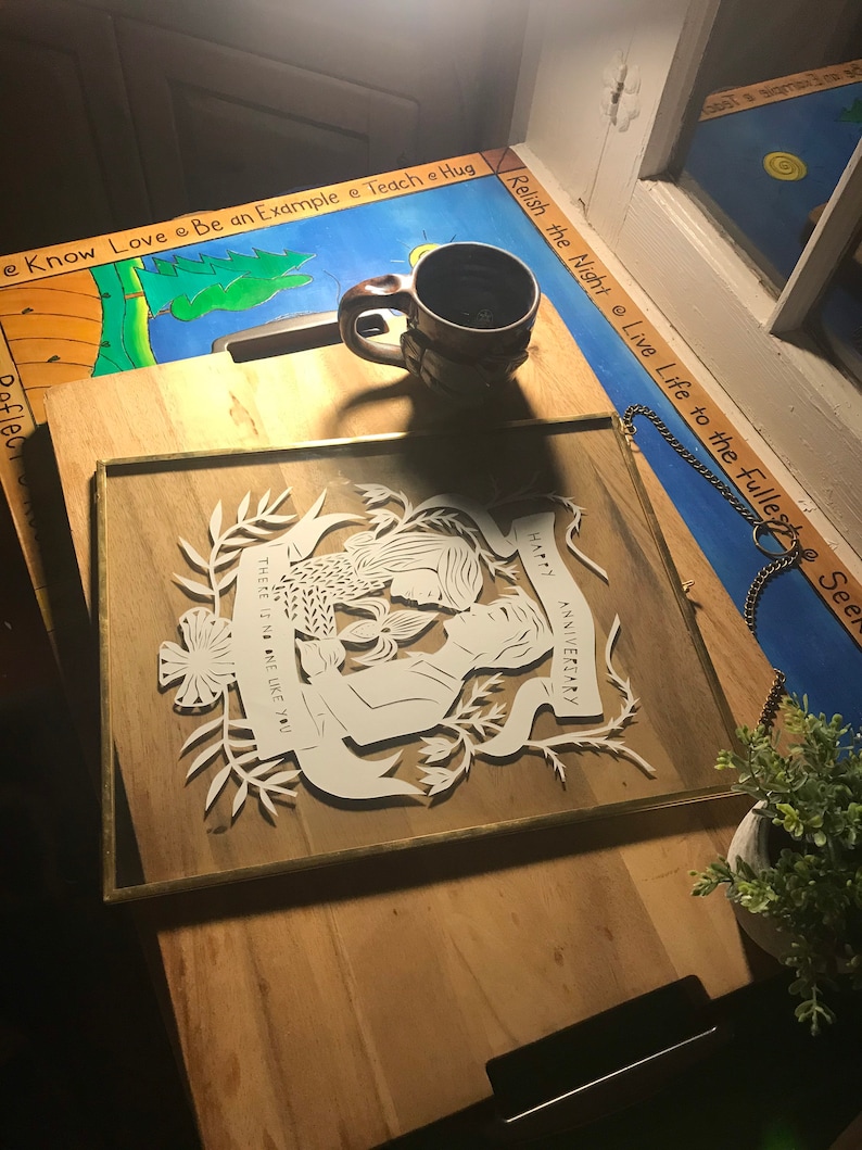 May include: A framed paper cut-out with a wood backing. The white cut-out features a couple embracing, floral elements, and the words "HAPPY ANNIVERSARY" and "THERE IS NO ONE LIKE YOU". A brown mug sits on a wooden surface.