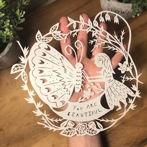 May include: White paper cut-out art featuring a butterfly, a girl, and flowers. The art is framed with a vine and the text "You are beautiful" is displayed on a banner.