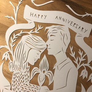 May include: A white paper cut-out art piece on a wooden surface. The design features a couple embracing, with floral elements and a banner that reads "HAPPY ANNIVERSARY." The artwork is intricate and detailed, with delicate cut lines.