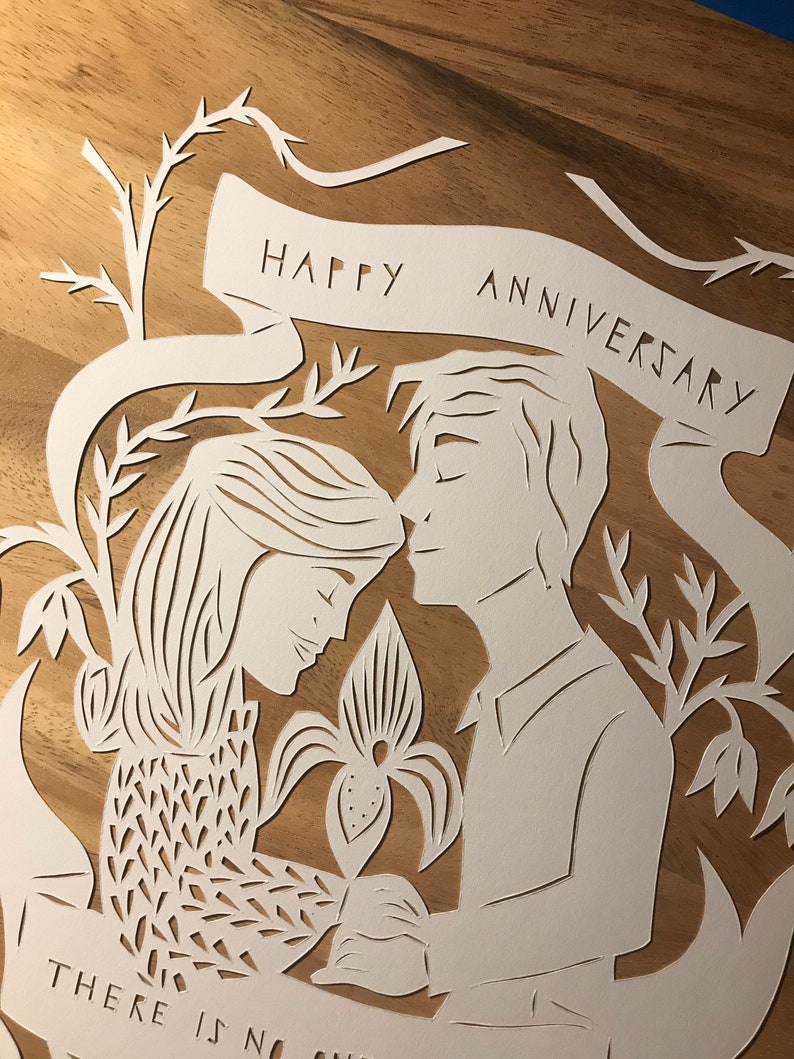 May include: A white paper cut-out art piece on a wooden surface. The design features a couple embracing, with floral elements and a banner that reads "HAPPY ANNIVERSARY." The artwork is intricate and detailed, with delicate cut lines.