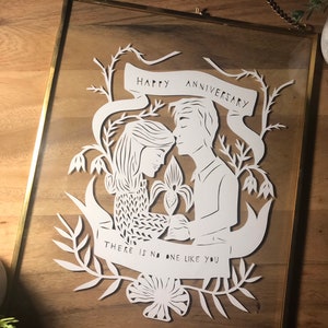May include: A framed paper cut art piece with a gold frame. The white paper cut depicts a couple with the text "HAPPY ANNIVERSARY" and "THERE IS NO ONE LIKE YOU". The art is surrounded by floral and leaf designs.