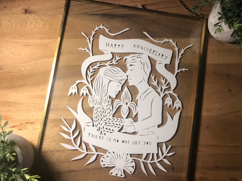 May include: A framed paper cut art piece with a gold frame. The white paper cut depicts a couple with the text "HAPPY ANNIVERSARY" and "THERE IS NO ONE LIKE YOU". The art is surrounded by floral and leaf designs.