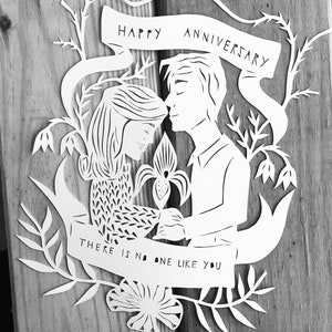 May include: A white paper cut-out anniversary card featuring a couple embracing, surrounded by floral and leaf designs. The banner at the top reads "HAPPY ANNIVERSARY," and the bottom banner says "THERE IS NO ONE LIKE YOU."