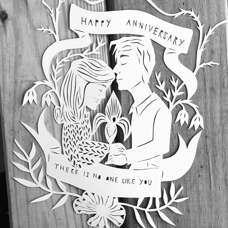 May include: A white paper cut-out anniversary card featuring a couple embracing, surrounded by floral and leaf designs. The banner at the top reads "HAPPY ANNIVERSARY," and the bottom banner says "THERE IS NO ONE LIKE YOU."