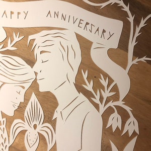 May include: A white paper cut-out art piece with the words "HAPPY ANNIVERSARY" on a banner. The artwork depicts a couple in profile, with floral and botanical elements. The background is a warm, wooden surface.