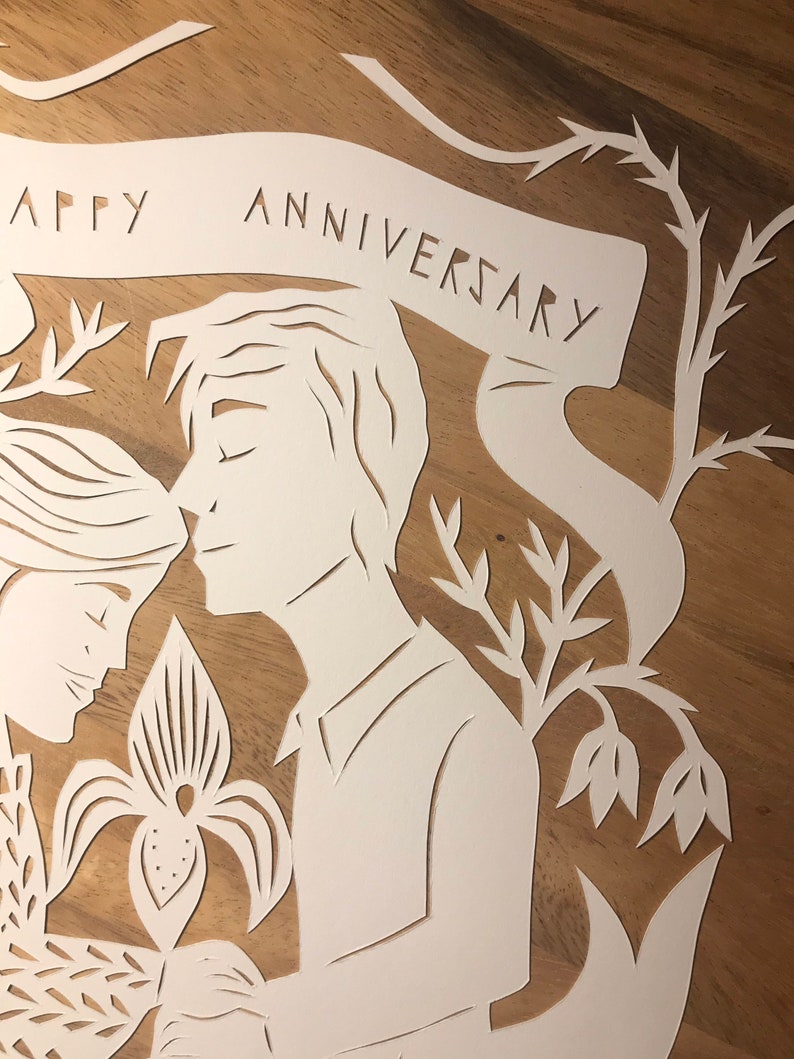 May include: A white paper cut-out art piece with the words "HAPPY ANNIVERSARY" on a banner. The artwork depicts a couple in profile, with floral and botanical elements. The background is a warm, wooden surface.