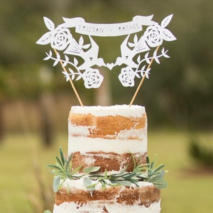May include: A white paper cake topper with two birds, flowers, and a banner that reads "Tristan + Rachel". The topper is on top of a white and brown cake.