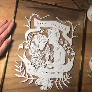 May include: White paper cut art in a gold frame. The art features a couple with the words "Happy Anniversary" and "There is no one like you". The art is surrounded by floral elements.