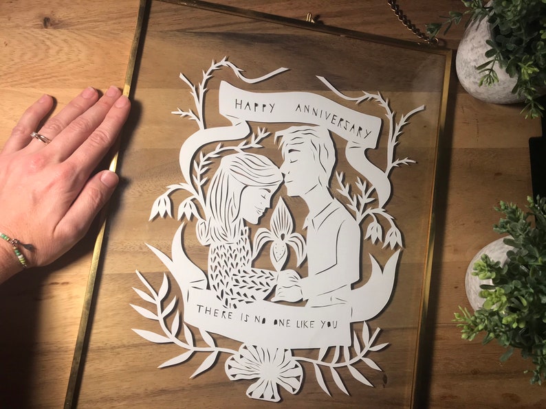 May include: White paper cut art in a gold frame. The art features a couple with the words "Happy Anniversary" and "There is no one like you". The art is surrounded by floral elements.