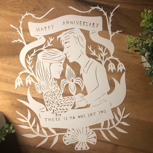 May include: A white paper cut-out of a couple with the words "HAPPY ANNIVERSARY" and "THERE IS NO ONE LIKE YOU". The artwork features floral and leaf designs, with the couple in a loving embrace. The background is a wooden surface.
