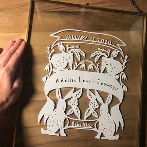 May include: A white paper cut-out art piece framed in gold. The art features two rabbits, flowers, and a banner that reads "Addison Lovett Connely's" and "January 15, 2020". The bottom of the banner reads "8lbs 10oz".
