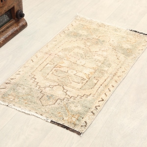 May include: Rectangular, vintage-style rug with a faded, distressed appearance. The rug features a central geometric design in shades of beige, brown, and green. The edges have a fringed detail. The rug is placed on a light-colored wooden floor.