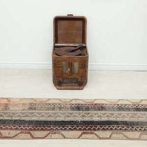 Puede incluir: Una alfombra rectangular de estilo vintage con un patrón geométrico en tonos beige, negro y rojo. La alfombra está colocada sobre un suelo claro. Una radio de madera vintage y un jarrón decorativo están en el fondo.