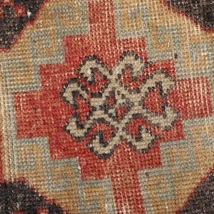 Puede incluir: Primer plano de una sección de una alfombra vintage con un patrón geométrico. La alfombra presenta un diseño central en rojo, negro y beige, rodeado por un patrón repetitivo en colores similares. La textura de la alfombra es visible.