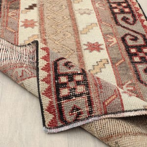 Può includere: Un tappeto rettangolare, tessuto a mano, con un angolo piegato, che mostra una miscela di tonalità beige, rosse e grigie. Il tappeto presenta un motivo a righe con motivi geometrici e floreali. I bordi sono rifiniti con una frangia.