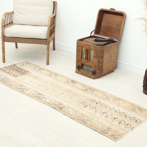 Op de afbeelding: Een rechthoekig, beige en bruin patroon tapijt met geometrische ontwerpen. Het tapijt ligt op een lichtgekleurde vloer, met een rieten stoel, een vintage houten radio en een decoratieve vaas op de achtergrond.