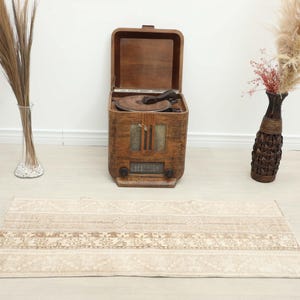 Könnte beinhalten: Ein beige-brauner gemusterter Teppich liegt auf einem hellen Holzboden. Hinter dem Teppich steht ein Vintage-Holzradio mit offenem Deckel. Hohe Vasen mit getrockneten Pflanzen stehen auf beiden Seiten des Radios.