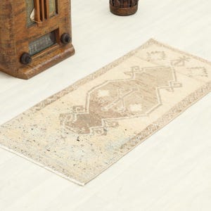 May include: A rectangular area rug featuring a central geometric medallion in shades of beige, cream, and light brown. The rug displays a symmetrical pattern with intricate borders and a weathered, distressed texture throughout the woven surface.