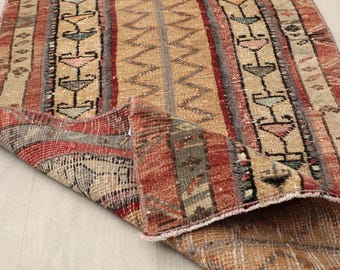 May include: A folded, rectangular rug with a distressed, vintage appearance. The rug features vertical stripes in shades of red, beige, and gray, with geometric patterns and a central zigzag design. The rug's edges are frayed, adding to its aged aesthetic.