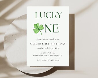 May include: A white invitation card with green text and a green shamrock illustration. The text reads "Lucky One" and "Oliver's 1st Birthday". The date and time of the party are listed as "March 18th 2 to 4 PM". The location is "At Oliver's Home 1234 Harbor Drive, San Diego, CA". The RSVP information is "RSVP to Jen 123-456-7890".