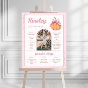 May include: A pink poster on an easel with the name "Kinsley" at the top. The poster includes a photo, a pumpkin illustration, and text detailing height, weight, favorite things, and nicknames. The height is 45.7 cm and the weight is 11.3 kg.