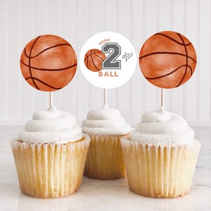 May include: Three cupcakes topped with basketball-themed paper toppers. Two toppers feature orange basketball illustrations, while the center topper is a white circle with a small basketball, the number 2, and the text BORN 2 BALL.