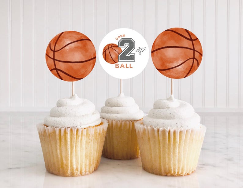 May include: Three cupcakes topped with basketball-themed paper toppers. Two toppers feature orange basketball illustrations, while the center topper is a white circle with a small basketball, the number 2, and the text BORN 2 BALL.