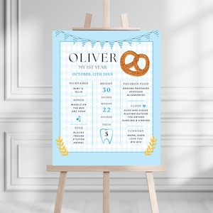 Può includere: Un poster azzurro su un cavalletto con la scritta "OLIVER MY 1ST YEAR OCTOBER, 12TH 20XX". Il poster include un pretzel, soprannomi, altezza 76 cm, peso 10 kg, cibi preferiti e altro.