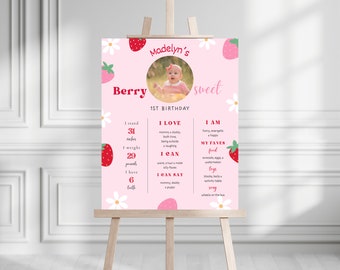 May include: A pink and white first birthday sign with a strawberry theme. The sign features a photo of a baby girl, along with details about her first year, including her height, weight, and favorite foods and activities. The text on the sign reads "Berry Sweet" and "Madelyn's 1st Birthday".