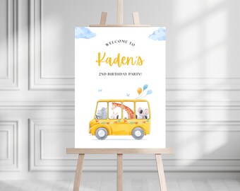 May include: Welcome to Kaden's 2nd Birthday Party! A yellow school bus with a giraffe, hippopotamus, rabbit, and koala riding inside. The bus has blue and yellow bunting and blue and yellow balloons.