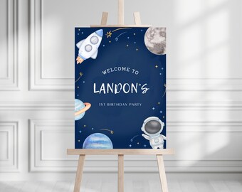 May include: A blue and white welcome sign with a space theme. The sign says "Welcome to Landon's 1st Birthday Party". The sign features a rocket, astronaut, planets, and stars.