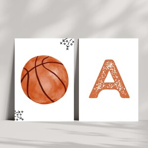 May include: Two rectangular white prints. The left print features a watercolor-style orange basketball with black seams and small black tactical play diagrams in the corners. The right print displays a large, textured orange letter A.