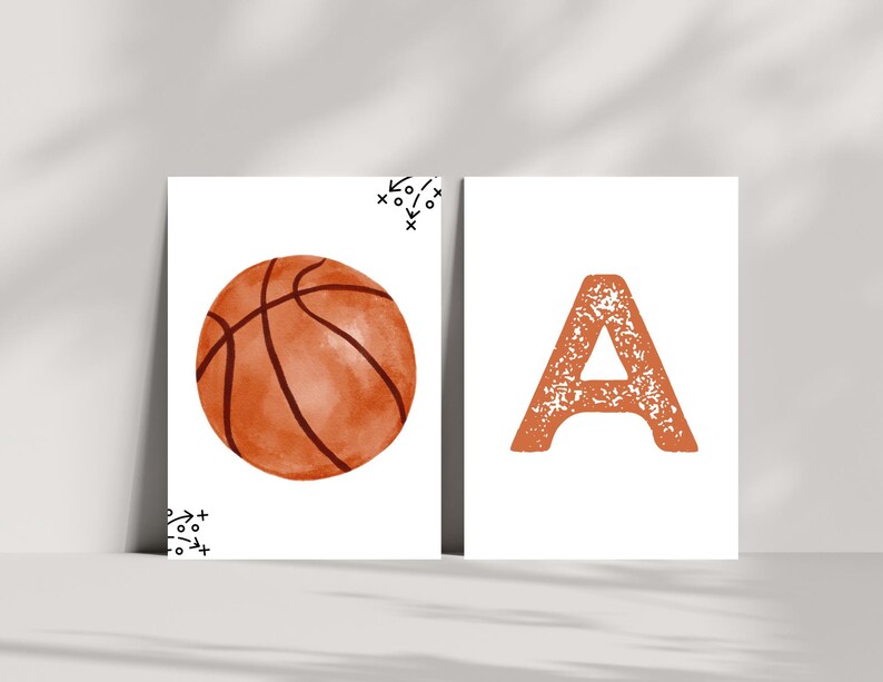 May include: Two rectangular white prints. The left print features a watercolor-style orange basketball with black seams and small black tactical play diagrams in the corners. The right print displays a large, textured orange letter A.