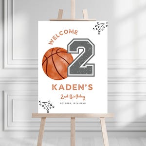 May include: A white rectangular birthday welcome sign featuring a basketball graphic, a large grey number 2, and the text WELCOME, KADEN'S, 2nd Birthday, and OCTOBER, 15TH 20XX in copper-colored lettering, accented with black sports play diagrams.