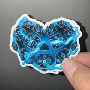 May include: A heart-shaped sticker featuring a design of blue and gold dice, with a lightning bolt effect. The dice have symbols on them. The sticker is held by a hand.