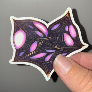 May include: A sticker with a black and gold design featuring a pattern of pink and purple shapes. The shapes are abstract and resemble eyes or flowers.