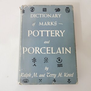 May include: A vintage book titled "Dictionary of Marks - Pottery and Porcelain" by Ralph M. and Terry H. Kovel. The book has a light blue cover with white text and various black illustrations. The cover shows signs of wear.