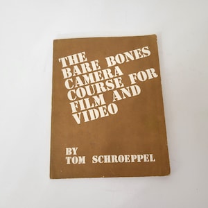 May include: A vintage book titled "The Bare Bones Camera Course for Film and Video" by Tom Schroeppel. The cover is a solid brown color with white text. The book appears to be in good condition, with slight wear on the edges.