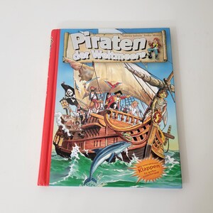 May include: A children's book titled "Pirates of the Seas" with a red spine. The cover features a detailed illustration of a pirate ship with pirates, a dolphin, and the sea. The book has German text.