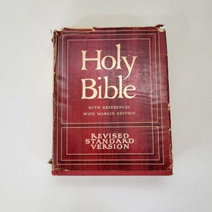 May include: A vintage Holy Bible with a burgundy cover. The title "Holy Bible" is prominently displayed in large, white lettering. The text "Revised Standard Version" is also visible. The book has a wide margin edition.