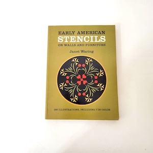 May include: A softcover book titled Early American Stencils on Walls and Furniture by Janet Waring. The olive green cover features a central black circular emblem with yellow and red floral patterns and text in white and black.
