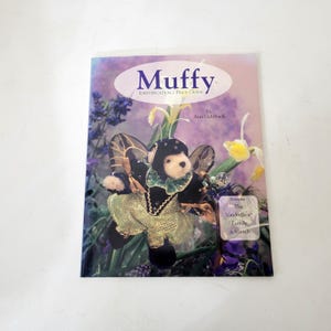May include: A book titled "Muffy Identification & Price Guide" featuring a teddy bear dressed as a fairy. The cover has a floral background with the text "by Ann Gehlbach" and "The VanderBear Family & friends".