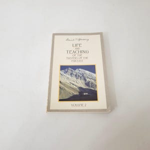 May include: A softcover book titled Life and Teaching of the Masters of the Far East Volume 2 by Baird T. Spalding. The cover features a rectangular image of snow-capped mountains and a small building against a clear blue sky.