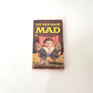 May include: Vintage MAD Magazine cover titled "The Self-Made MAD." The cover features a cartoon illustration of Alfred E. Neuman with his feet up on a desk, smoking a cigar. The magazine is brown and yellow.